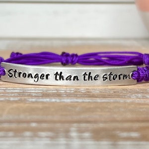 Op de afbeelding: Paarse gevlochten armband met een zilveren bedel met de tekst "Sterker dan de storm".