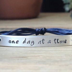 May include: A silver bracelet with a navy blue cord. The bracelet has a silver tag with the words "one day at a time" engraved on it.