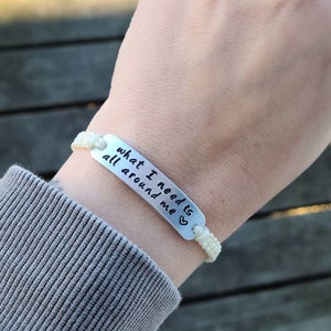 Puede incluir: Pulsera trenzada blanca con un dije plateado que dice "what I need is all around me" con un peque&ntilde;o s&iacute;mbolo de coraz&oacute;n.