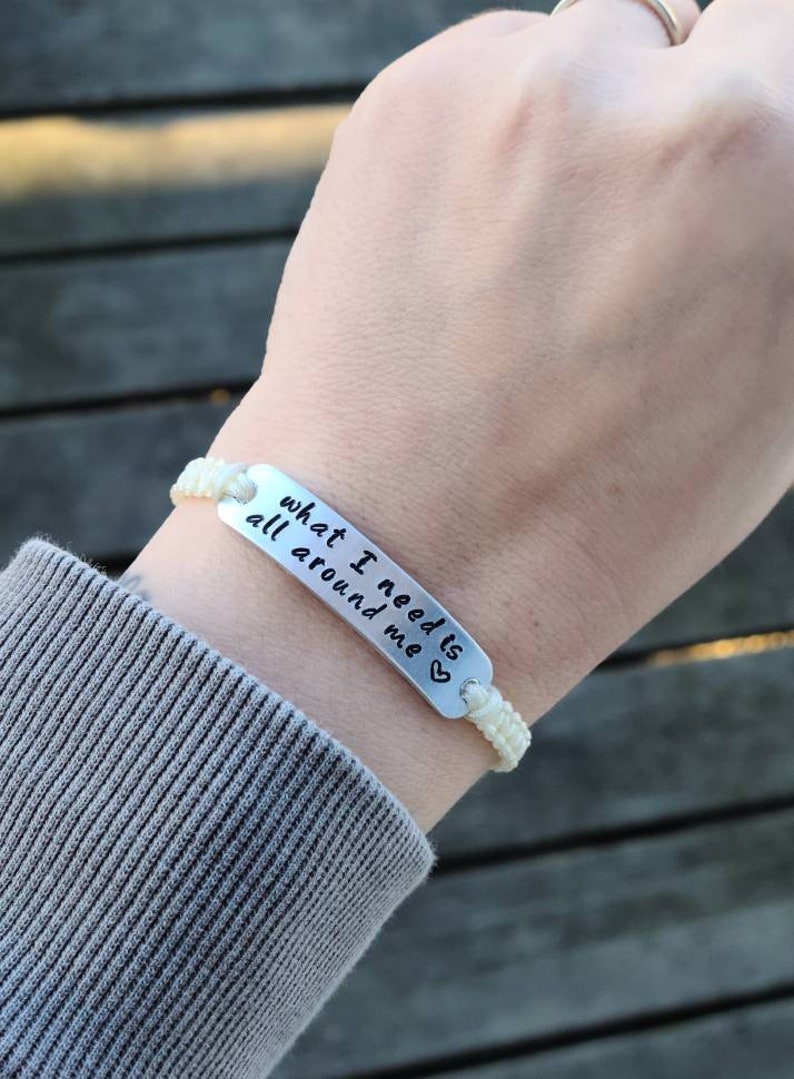 Puede incluir: Pulsera trenzada blanca con un dije plateado que dice "what I need is all around me" con un peque&ntilde;o s&iacute;mbolo de coraz&oacute;n.