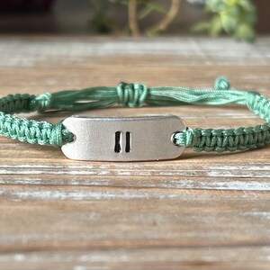 Op de afbeelding: Handgemaakte macram&eacute; armband in een zachte groene tint. De armband heeft een geweven ontwerp en een rechthoekige zilverkleurige metalen plaat met twee verticale lijnen. De armband ligt op een houten oppervlak.