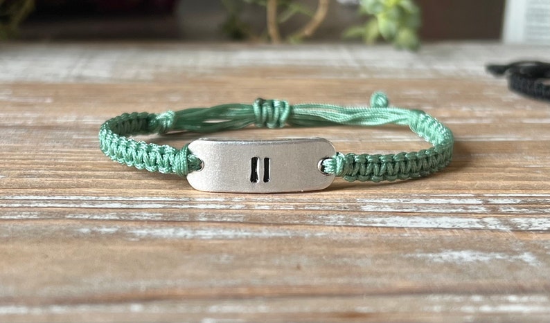 Op de afbeelding: Handgemaakte macram&eacute; armband in een zachte groene tint. De armband heeft een geweven ontwerp en een rechthoekige zilverkleurige metalen plaat met twee verticale lijnen. De armband ligt op een houten oppervlak.