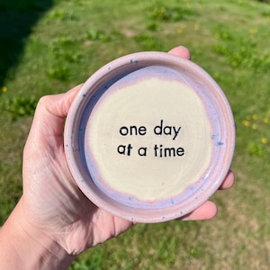 May include: A round ceramic trinket dish with a cream-colored center and a light purple rim. The dish features the black text "one day at a time" stamped in the middle. The surface has a speckled texture and a smooth, glossy finish throughout.