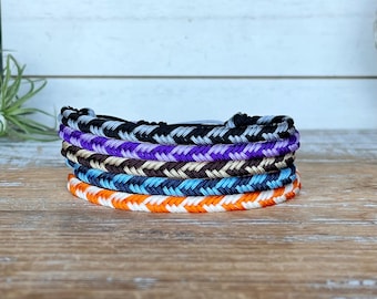 May include: A stack of six braided friendship bracelets in various colors, including black, purple, brown, blue, and orange. The bracelets are made of woven thread and rest on a wooden surface.
