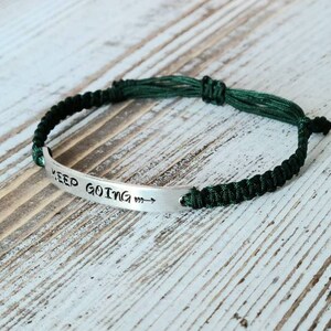 May include: A silver bracelet with the words "KEEP GOING" stamped on a curved metal plate. The bracelet is attached to a dark green braided cord. The arrow points to the right.