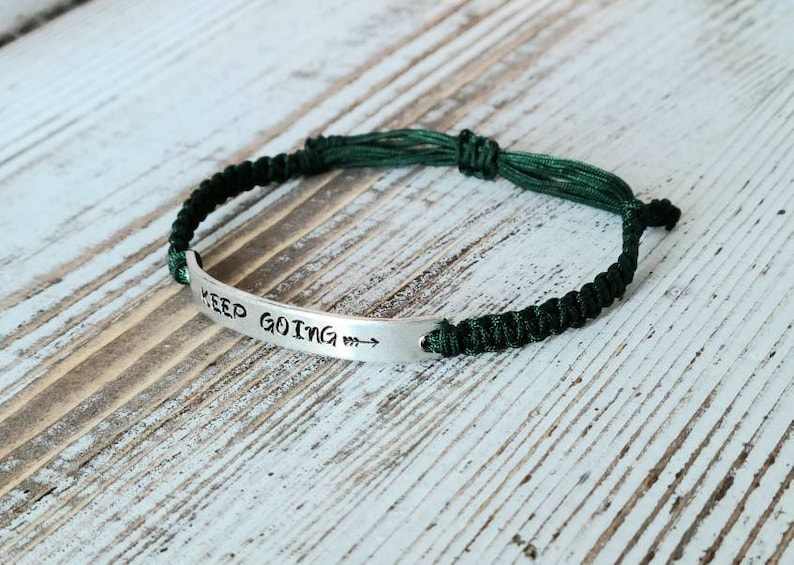 May include: A silver bracelet with the words "KEEP GOING" stamped on a curved metal plate. The bracelet is attached to a dark green braided cord. The arrow points to the right.