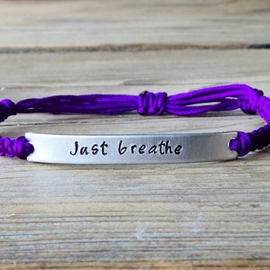 Op de afbeelding: Een armband van paars gevlochten koord met een zilveren staafbedel met de tekst "Just breathe".