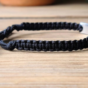 May include: Black braided bracelet with a silver metal tag. The tag has an inscription.