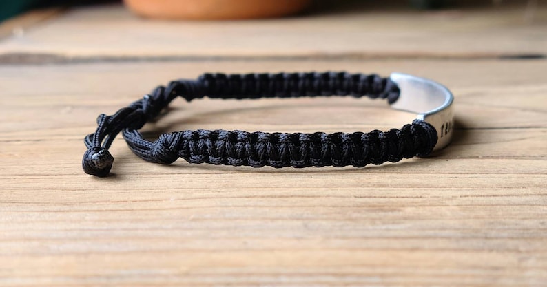 May include: Black braided bracelet with a silver metal tag. The tag has an inscription.