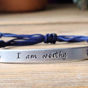 May include: A navy blue braided bracelet with a silver metal charm that reads "I am worthy".