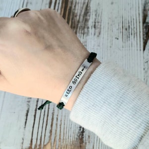May include: A silver bracelet with the words "KEEP GOING" and an arrow symbol. The bracelet is attached to a black cord and green knot. The bracelet is worn on a wrist.