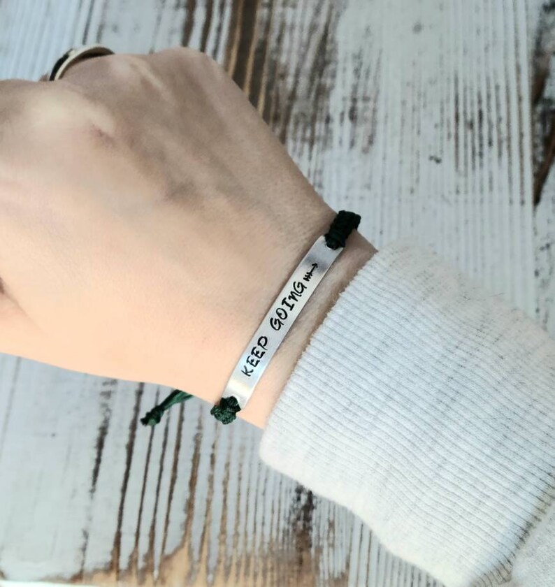 May include: A silver bracelet with the words "KEEP GOING" and an arrow symbol. The bracelet is attached to a black cord and green knot. The bracelet is worn on a wrist.