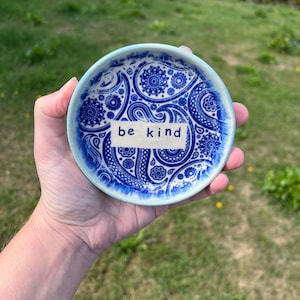 May include: A small, round ceramic trinket dish featuring a vibrant blue and white paisley pattern. The center of the dish displays a rectangular white label with the text "be kind" printed in a simple, dark blue sans-serif font.