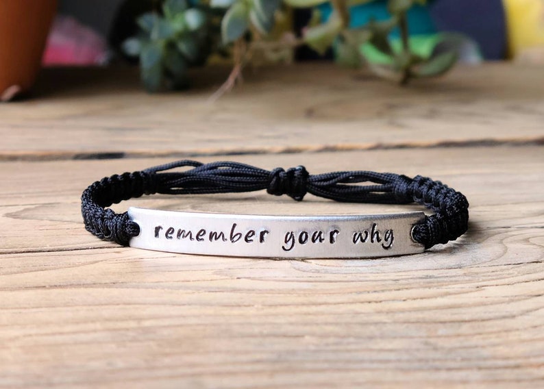 May include: A black cord bracelet with a silver metal charm that reads "remember your why".