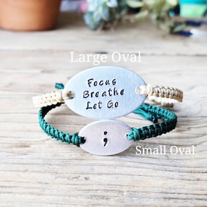 Puede incluir: Dos pulseras de amuleto ovaladas de plata con cordones verdes y beige. El amuleto m&aacute;s grande dice "Focus Breathe Let Go" y el amuleto m&aacute;s peque&ntilde;o tiene un punto y coma.