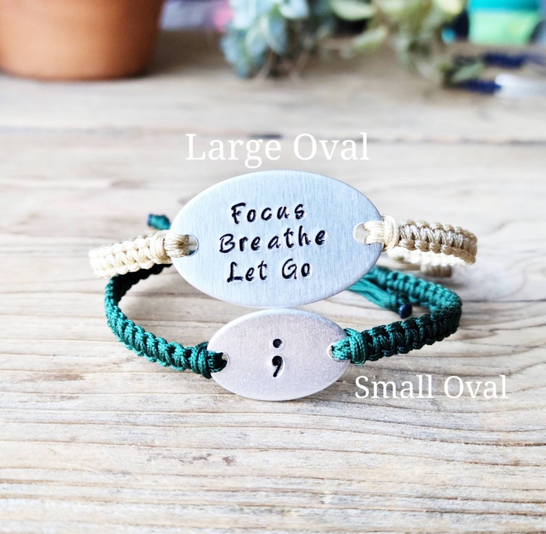 Puede incluir: Dos pulseras de amuleto ovaladas de plata con cordones verdes y beige. El amuleto m&aacute;s grande dice "Focus Breathe Let Go" y el amuleto m&aacute;s peque&ntilde;o tiene un punto y coma.