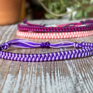 May include: A purple and light purple braided friendship bracelet with a tassel. The bracelet is made with a chevron pattern.