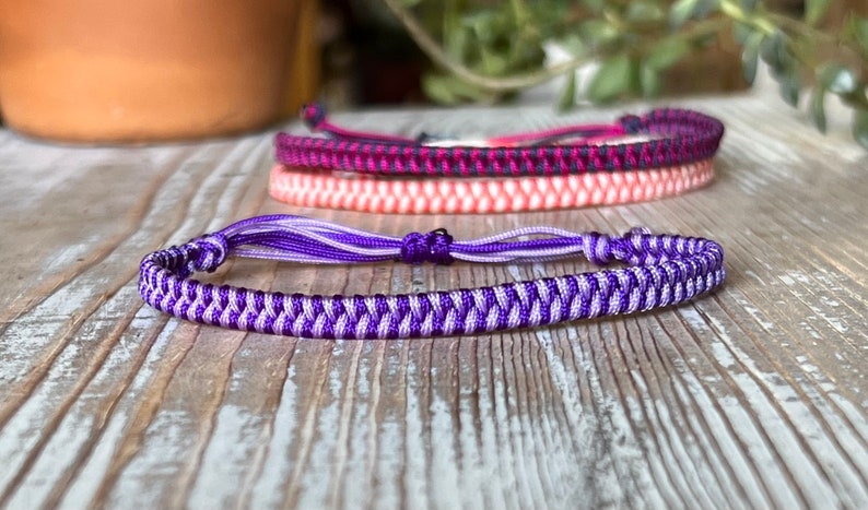 May include: A purple and light purple braided friendship bracelet with a tassel. The bracelet is made with a chevron pattern.