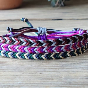 May include: A set of four braided friendship bracelets in various colors, including green, black, white, pink, and purple. The bracelets are woven together in a herringbone pattern.
