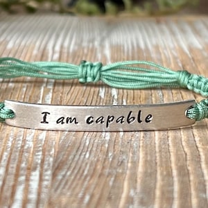 May include: A green braided cord bracelet with a silver charm that reads "I am capable".