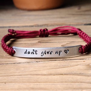 Op de afbeelding: Een armband van gevlochten maroonkleurig koord met een zilveren bedel waarop "geef nooit op" staat met een klein hartje.