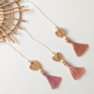 Op de afbeelding: Drie geweven hartvormige ornamenten met houten kralen en kwastjes. De kwastjes zijn roze en oranje.