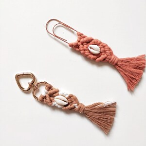 Op de afbeelding: Twee handgemaakte macramé sleutelhangers met een hartvormige metalen ring en een kwast. Elke sleutelhanger heeft een witte schelp accent.