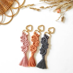 Puede incluir: Tres llaveros de macramé hechos a mano con anillas en forma de corazón. Los llaveros están hechos con hilo rosa, naranja y gris y tienen borlas en la parte inferior.