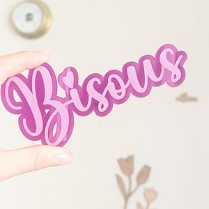 May include: A translucent, magenta-colored sign with the word "Bisous" in a cursive font. The sign is held up by a hand. The word "Bisous" is French for "kisses".