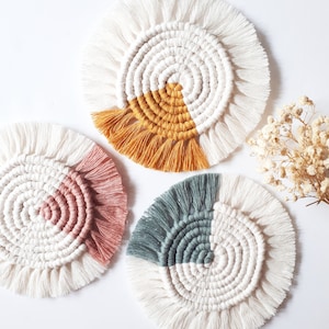 Op de afbeelding: Drie handgemaakte macramé-onderzetters met een wit centrum en een gekleurd accent. De onderzetters zijn rond en hebben een franje rand. De kleuren zijn mosterdgeel, stoffig roze en teal.