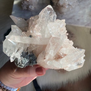 May include: A cluster of clear quartz crystals with a hint of pink, forming a large, intricate cluster. The crystals are translucent and have a natural, rough texture.