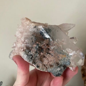 May include: A cluster of clear quartz crystals with a pink hue, embedded in a matrix of gray and brown rock. The crystals have a variety of shapes and sizes, and some have a pointed tip.