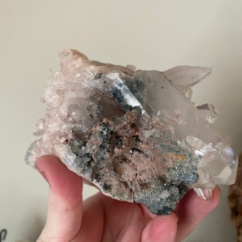 May include: A cluster of clear quartz crystals with a pink hue, embedded in a matrix of gray and brown rock. The crystals have a variety of shapes and sizes, and some have a pointed tip.