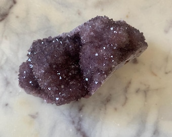 May include: A cluster of deep purple amethyst crystals. The crystals have a sparkling, textured surface and are clustered together on a rough, natural base. The amethyst is a rich, dark shade of purple.