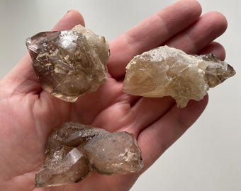 May include: Three smoky quartz crystals, each with a unique shape and size, are displayed on a hand. The crystals are translucent and have a light brown color.