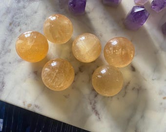 May include: A collection of polished, golden-yellow citrine spheres and faceted purple amethyst crystals. The spheres are arranged in the foreground, while the amethyst crystals are in the background. The crystals and spheres are on a light-colored surface.