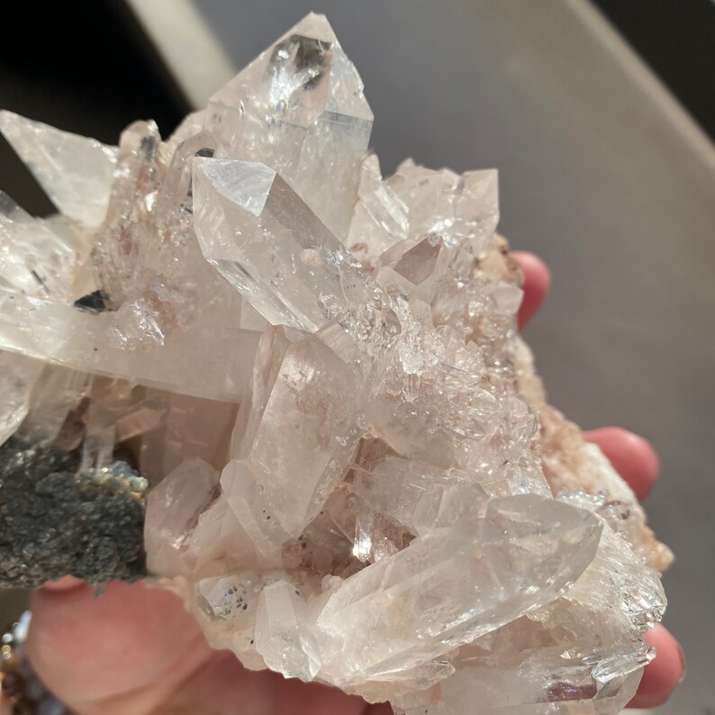 May include: A cluster of clear quartz crystals with a rough, natural base. The crystals are translucent and have a slight pink hue.