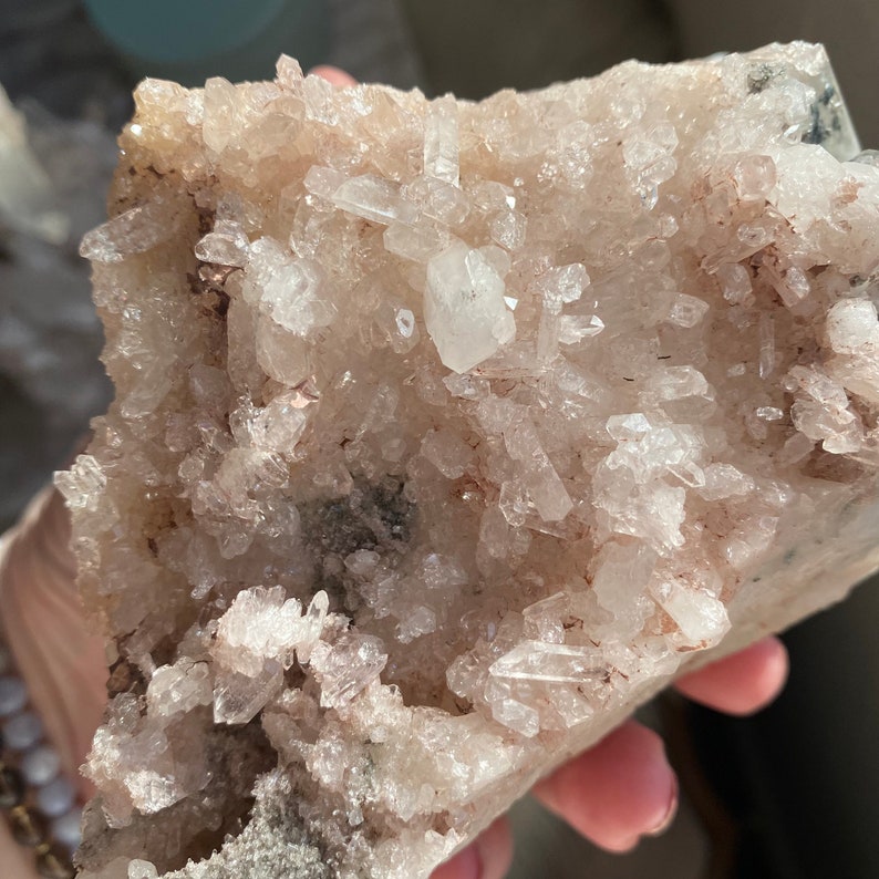 May include: A cluster of clear quartz crystals with a pink hue, embedded in a rough, gray rock. The crystals are of varying sizes and shapes, with some appearing to be elongated and pointed.