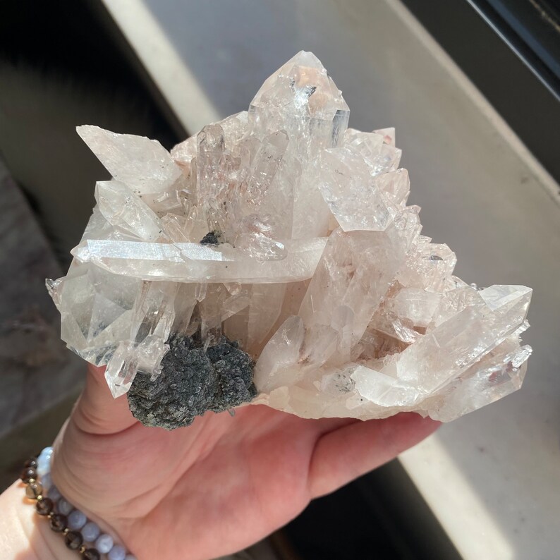 May include: A cluster of clear quartz crystals with a dark gray mineral base. The crystals are arranged in a pointed formation, with some of the crystals being longer than others.