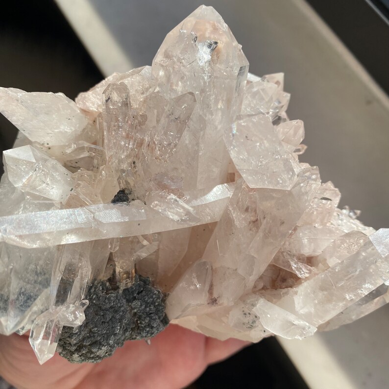 May include: A cluster of clear quartz crystals with a gray mineral base. The crystals are arranged in a cluster, with some pointing upwards and others pointing downwards. The crystals are clear and have a slight pink hue.