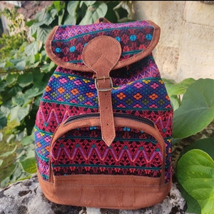 Op de afbeelding: Een kleurrijke rugzak met een geometrisch patroon in tinten roze, rood, blauw, groen en oranje. De rugzak heeft een bruine leren klep met een gesp en een ritsvak.