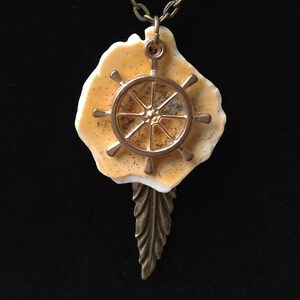 Pode incluir: Um colar com um pingente de roda de navio dourado, um charme em forma de concha bege e branca e um charme em forma de pena de bronze.