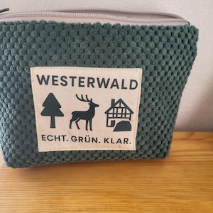 Könnte beinhalten: Eine dunkelgrüne, strukturierte Kosmetiktasche mit beigem Reißverschluss und einem rechteckigen Aufnäher. Der Aufnäher zeigt das Wort "WESTERWALD" über einer Baum-, Hirsch- und Hausillustration mit dem Text "ECHT. GRÜN. KLAR."