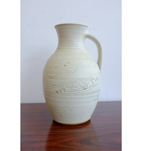 May include: A white ceramic vase with a textured design and a handle. The vase has a rounded body and a slightly flared opening.