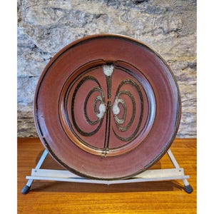 May include: A round, reddish-brown ceramic plate with a decorative black and white spiral design. The plate is displayed on a silver metal stand, showcasing its unique artistic pattern. The plate's rim is a darker shade of brown.