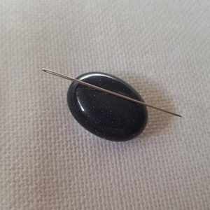 Op de afbeelding: Een donkerblauwe, ovale kraal met een naald erdoorheen.
