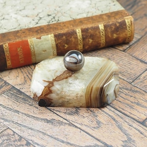 Puede incluir: Una piedra de ágata pulida con bandas marrones y blancas, coronada por una pequeña canica esférica en blanco y negro. Un libro antiguo con una cubierta de cuero marrón está en el fondo. Los objetos están sobre una superficie de madera.