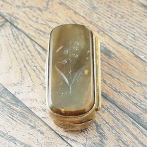 May include: A rectangular gold-toned metal box featuring a smooth, translucent brown stone inlay on the lid. The stone is engraved with a delicate floral design of a stem with small blossoms and leaves centred on the surface of the lid.