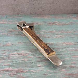 May include: A vintage bottle opener with a metal head and a deer antler handle. The handle is a mix of brown and cream colours. The opener is on a weathered, teal-coloured wooden surface.