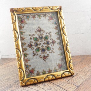 May include: A gold-coloured picture frame with a detailed, ornate border. The frame holds a piece of fabric with a colourful, embroidered floral pattern in shades of red, green, blue, and gold. The frame is angled on a wooden surface.